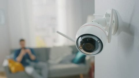 A white indoor security camera mounted on a wall in a brightly lit living room, demonstrating the kind of equipment used by a CCTV monitoring company for residential and home CCTV monitoring.