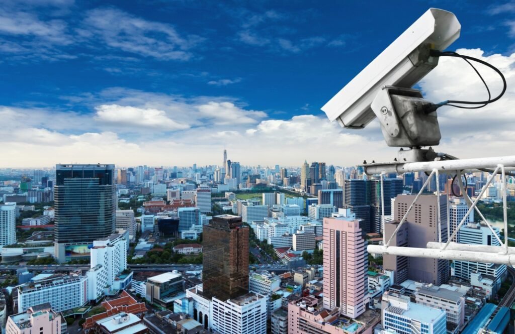 White bullet-style security camera mounted on a pole against a vibrant cityscape, symbolizing nationwide CCTV monitoring and urban business security.
