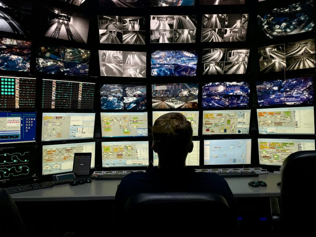Rear view of a person sitting in a control room, surrounded by dozens of large monitors displaying various real-time video feeds and complex system diagrams.