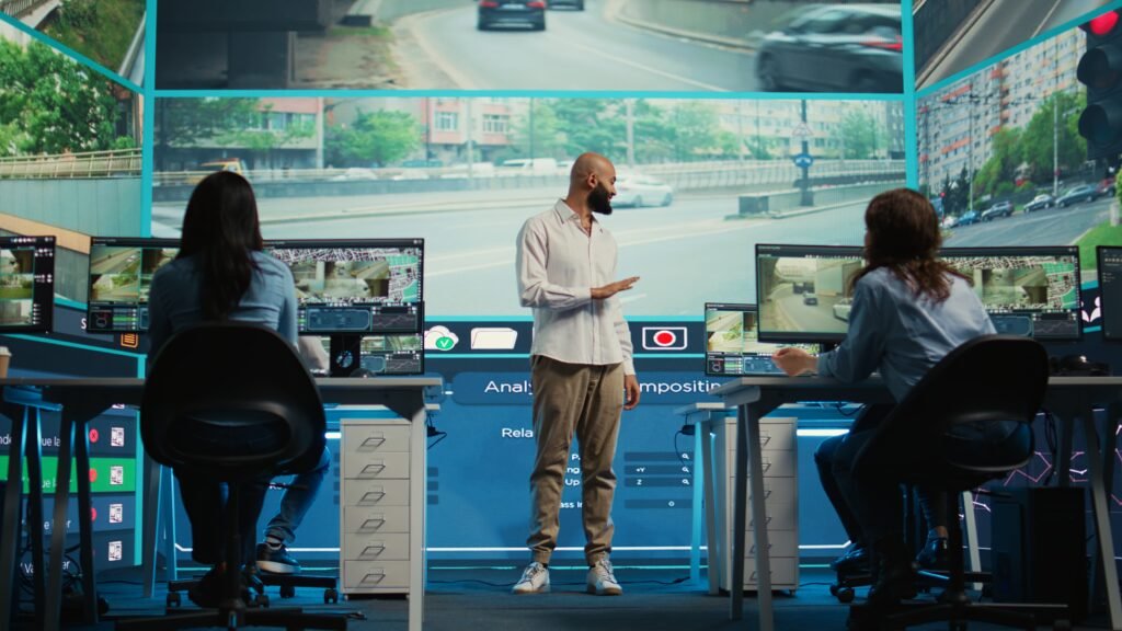 A team leader briefs two employees in a high-tech monitoring room with large screens displaying traffic, representing the structured operations of a dedicated CCTV monitoring company.