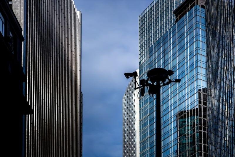 A silhouette of a multi-camera security pole set against modern glass skyscrapers, representing Trusted CCTV Monitoring Services and elite security cameras monitoring services in an urban environment.