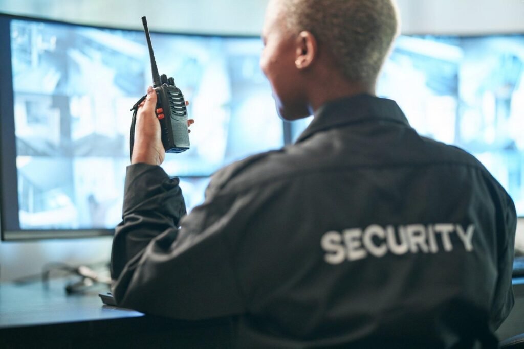 Alt Text: A professional security guard communicating via a walkie-talkie in front of a multi-screen camera console, illustrating the advanced business surveillance and commercial CCTV operations utilized by the Top Security Providers.