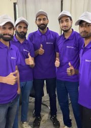 Five team members in purple GCCTVMS uniforms giving thumbs up in a security monitoring center, representing virtual doorman and virtual guard services.