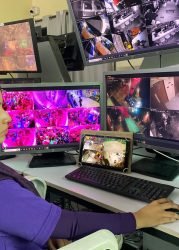 Female operator in GCCTVMS uniform monitoring live surveillance footage on several computer screens in a control room.