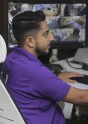 Male operator in a purple GCCTVMS shirt monitoring surveillance footage on a computer screen.