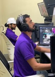 Male technicians in purple GCCTVMS uniforms wearing headsets and monitoring multiple camera feeds on computer screens.